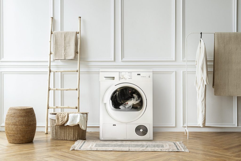 A white condenser dryer with laundry in the drum, standing against paneled walls beside a towel ladder