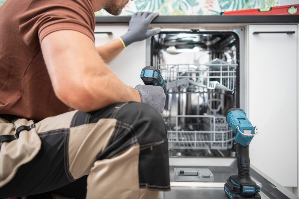 A man in work trousers kneeling at an open dishwasher, a cordless drill in the lower rack beside him